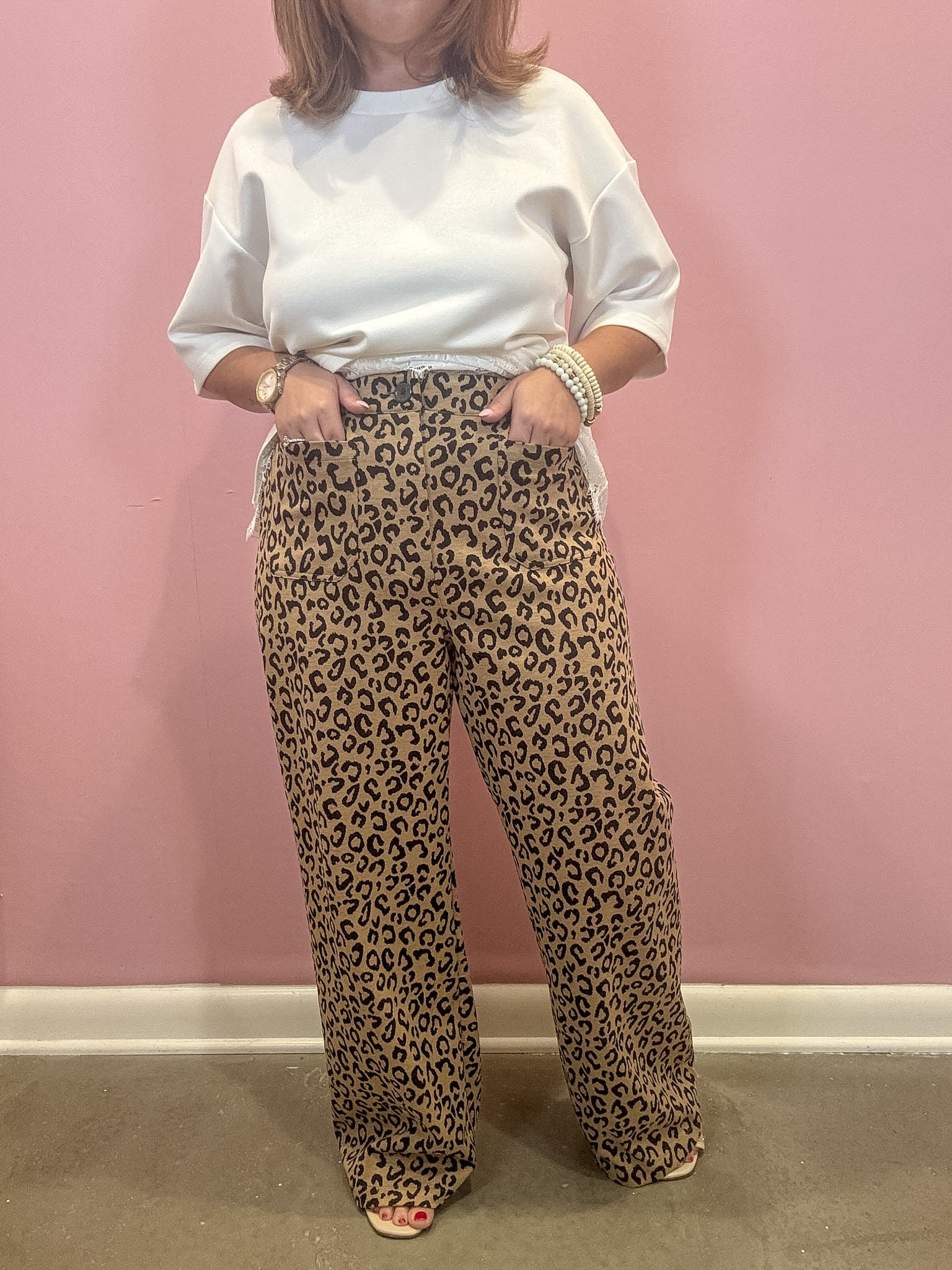 A person wearing loose leopard-print pants and a white sweatshirt stands against a pink wall, with hands in the pockets of the pants.