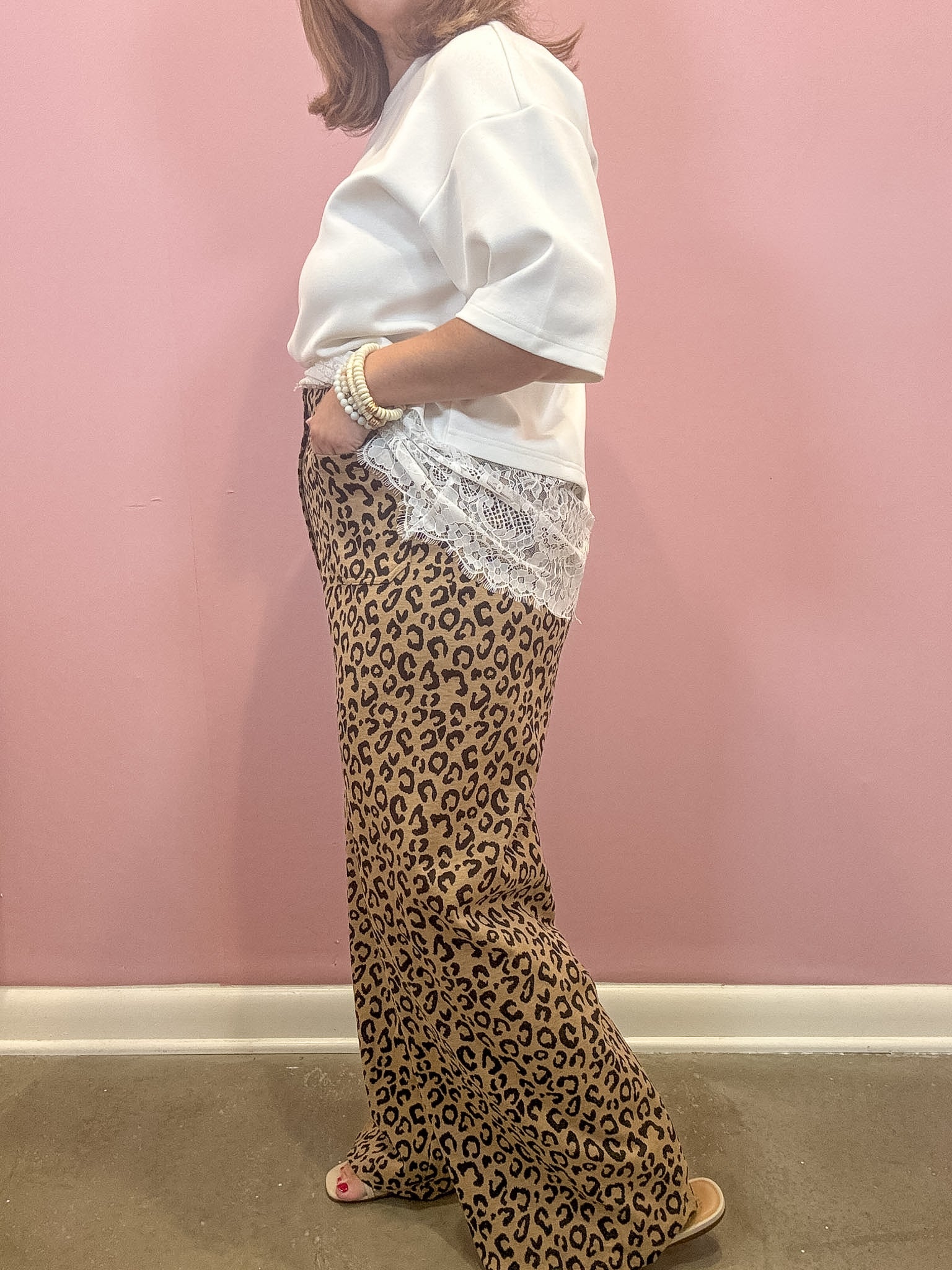 A woman wearing a white short-sleeve top and leopard-print wide-leg pants with a white lace hem, standing against a pink wall.