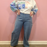 A person stands against a pink wall wearing a white graphic sweatshirt with blue and green patterned sleeves, light blue wide-leg jeans, and beige heeled sandals.