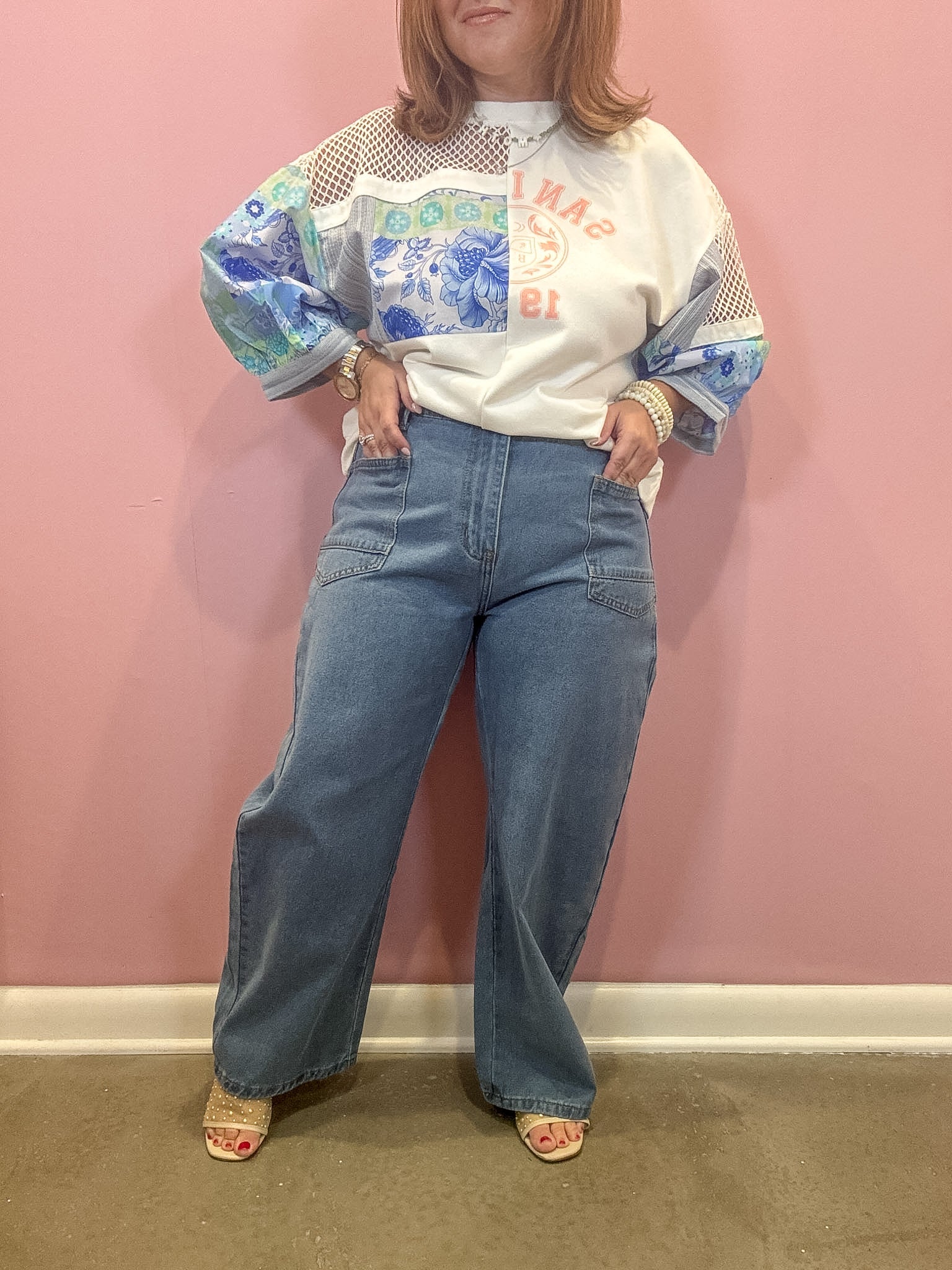 A person stands against a pink wall wearing a white graphic sweatshirt with blue and green patterned sleeves, light blue wide-leg jeans, and beige heeled sandals.
