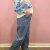 A person wearing a white long-sleeve blouse with blue floral and striped panels, paired with wide-leg blue jeans and beige slide sandals, standing against a pink wall.