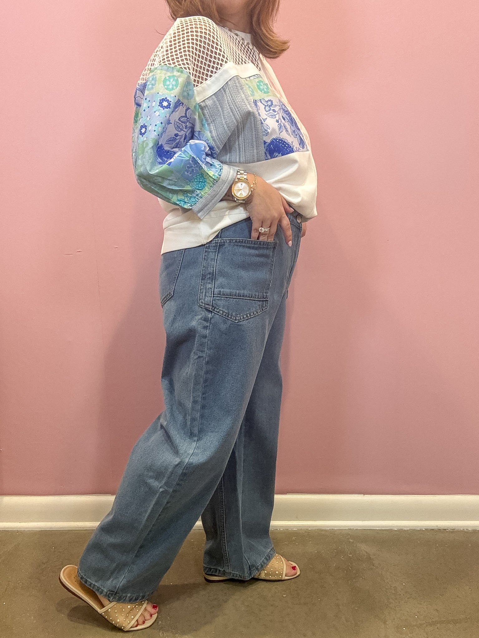 A person wearing a white long-sleeve blouse with blue floral and striped panels, paired with wide-leg blue jeans and beige slide sandals, standing against a pink wall.