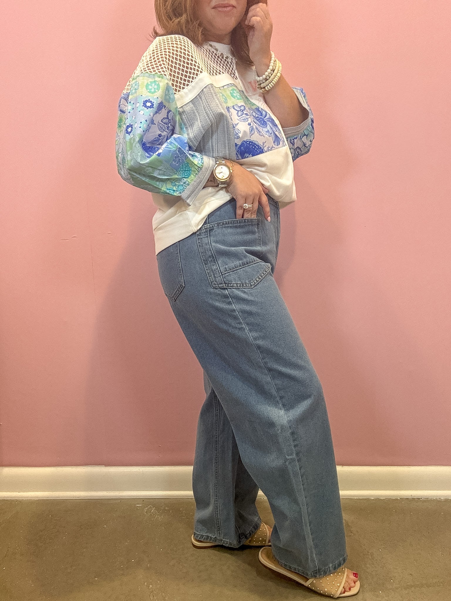 A woman modeling a white and blue patchwork blouse with floral long sleeves, paired with wide-leg blue jeans, standing against a pink wall.