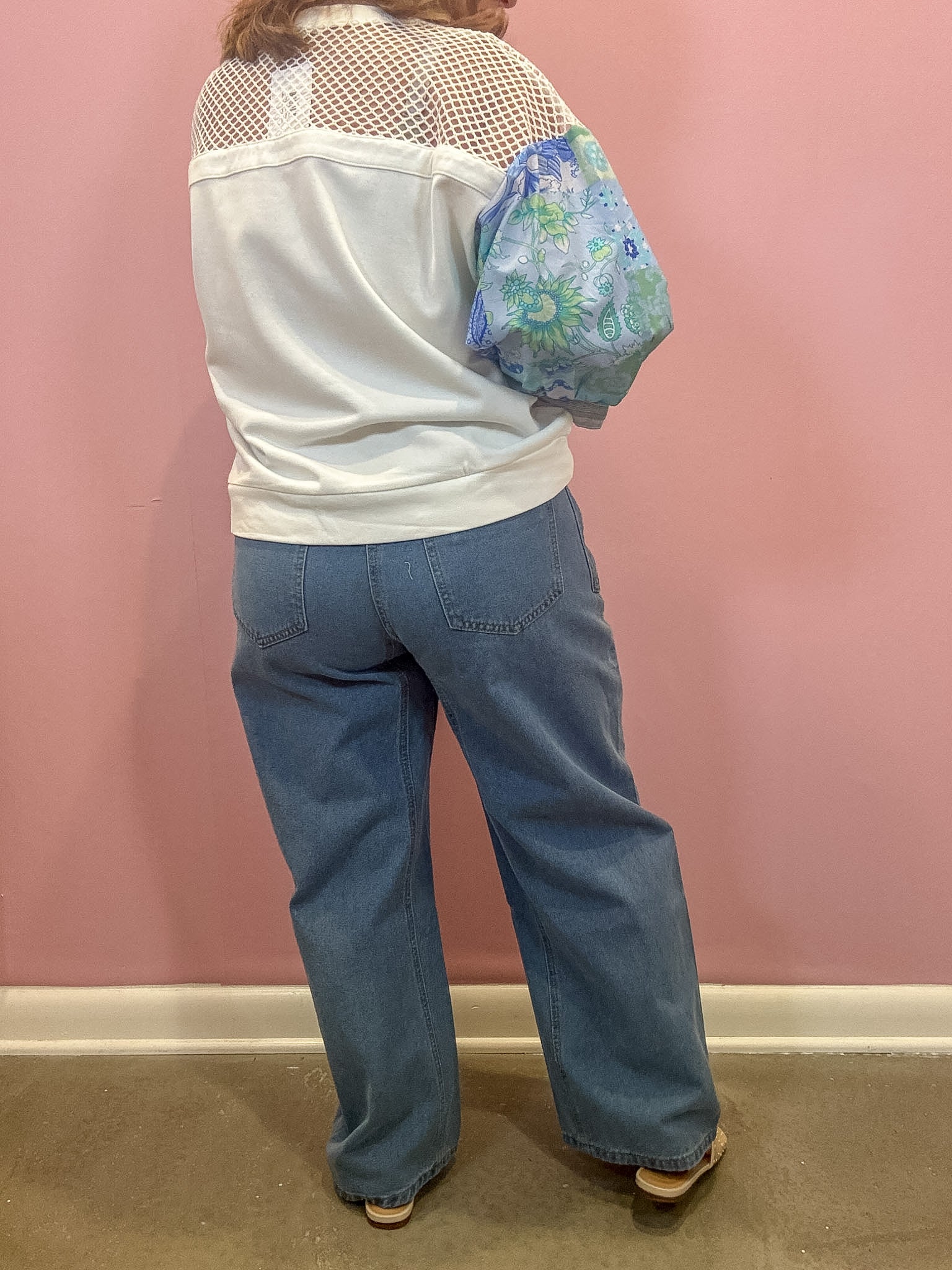 A person shown from the back wearing a white cropped top with a sheer mesh yoke, wide-leg blue jeans, and carrying a blue floral shoulder bag against a pink wall.