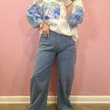 A woman modeling a white knit sweater with blue and green floral patterns on the sleeves and upper chest, paired with wide-leg blue jeans and beige heeled sandals, standing against a pink wall.
