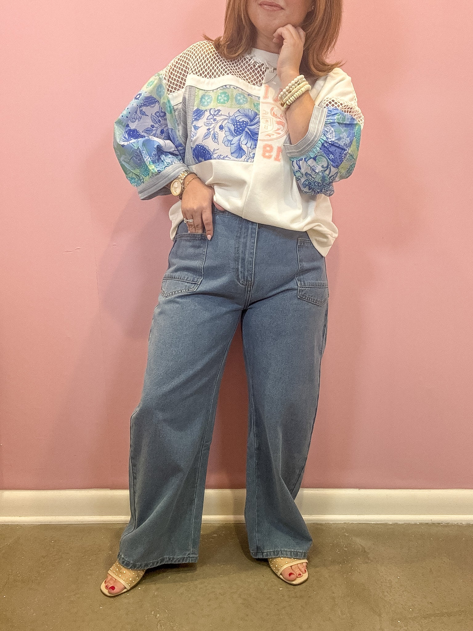 A woman modeling a white knit sweater with blue and green floral patterns on the sleeves and upper chest, paired with wide-leg blue jeans and beige heeled sandals, standing against a pink wall.