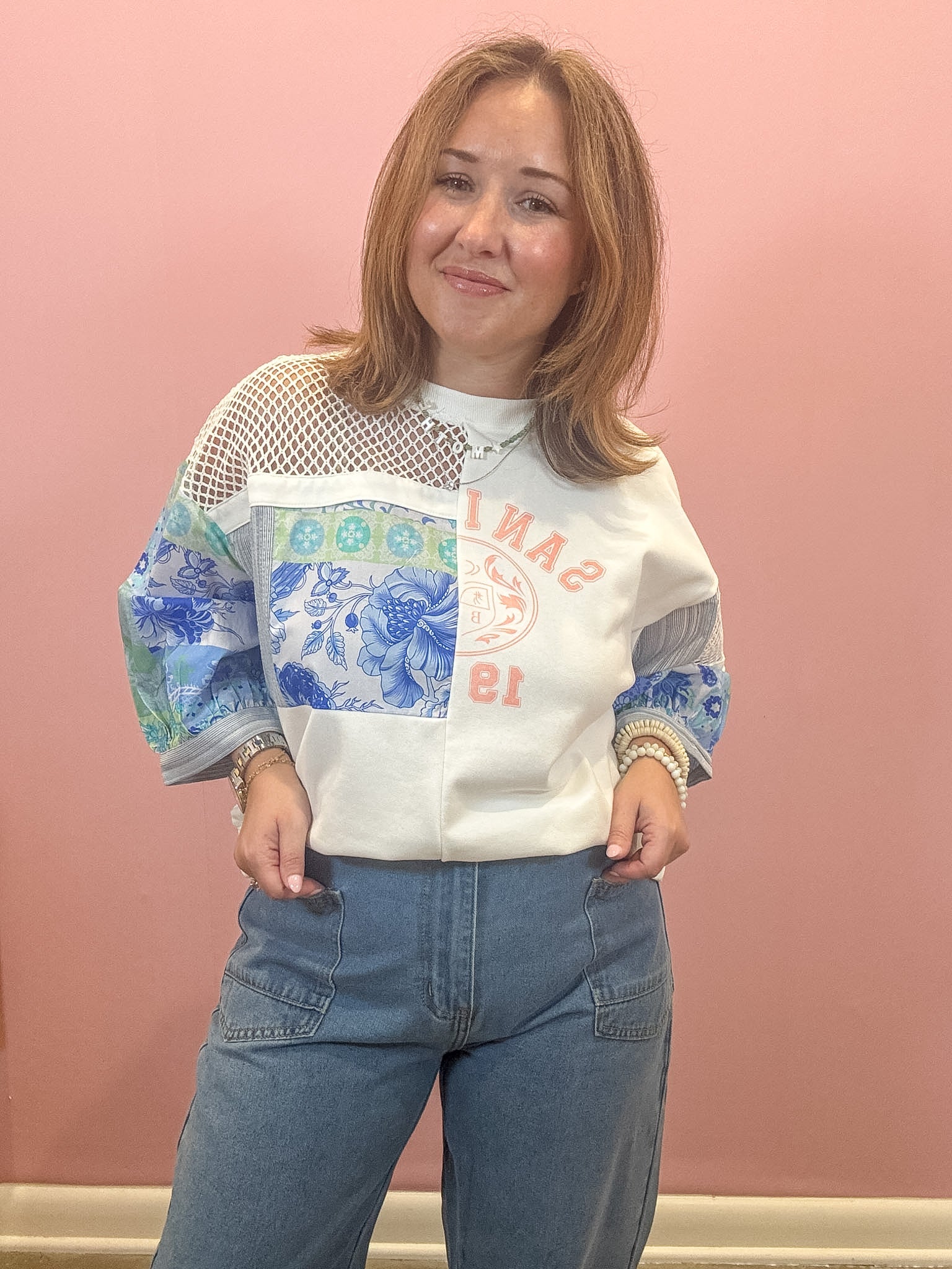 A woman modeling a white knit sweater with blue floral and geometric patterns on the sleeves and chest, paired with blue jeans, standing against a solid pink wall.