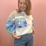 A woman modeling a white mock-neck blouse with blue floral and patterned patchwork sleeves, paired with blue jeans, standing against a solid pink wall.