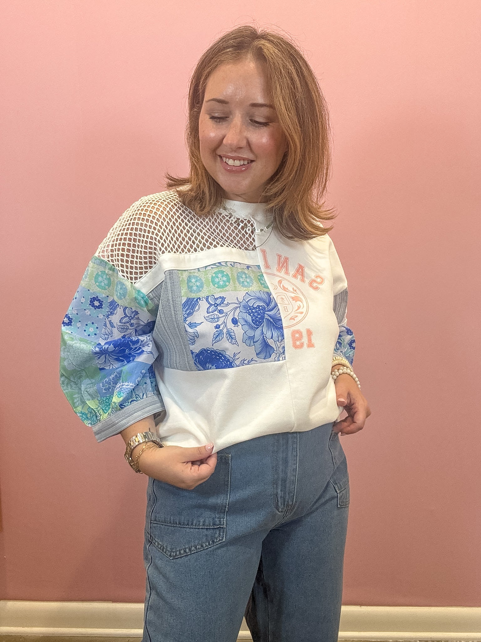 A woman modeling a white mock-neck blouse with blue floral and patterned patchwork sleeves, paired with blue jeans, standing against a solid pink wall.