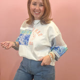 A woman modeling a white crewneck sweatshirt with blue floral panels and mesh sleeves, paired with blue jeans, standing against a solid pink wall.