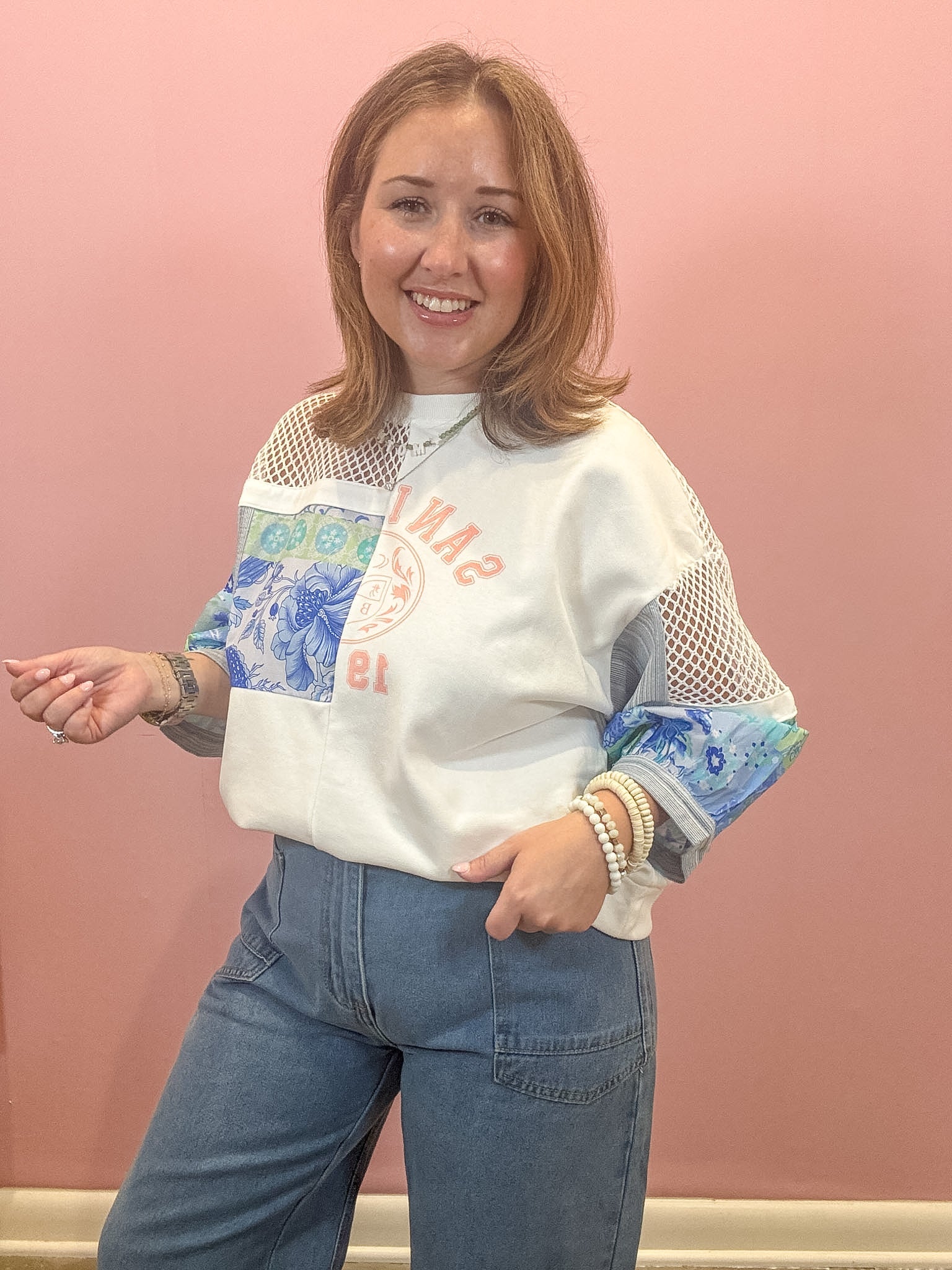 A woman modeling a white crewneck sweatshirt with blue floral panels and mesh sleeves, paired with blue jeans, standing against a solid pink wall.