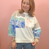 A woman wearing a white crewneck sweatshirt with blue and green patterned sleeves, mesh shoulder panels, and a red graphic print, paired with blue jeans, standing against a pink wall.