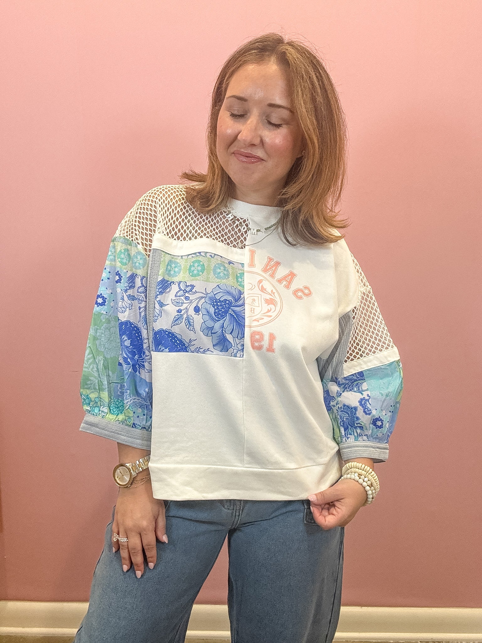 A woman wearing a white crewneck sweatshirt with blue and green patterned sleeves, mesh shoulder panels, and a red graphic print, paired with blue jeans, standing against a pink wall.