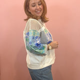 A woman wearing a white crewneck sweatshirt with sheer mesh sleeves and blue floral printed elbow-length sleeves, paired with blue jeans, standing against a solid pink wall.