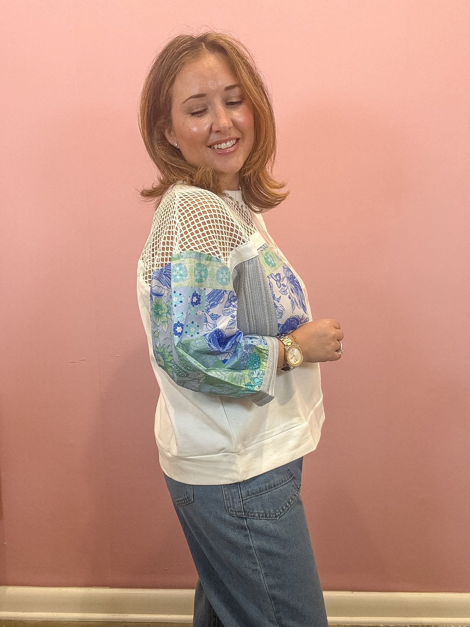A woman wearing a white crewneck sweatshirt with sheer mesh sleeves and blue floral printed elbow-length sleeves, paired with blue jeans, standing against a solid pink wall.