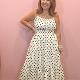 A woman modeling a white spaghetti-strap maxi dress with black polka dots, standing against a solid pink wall.
