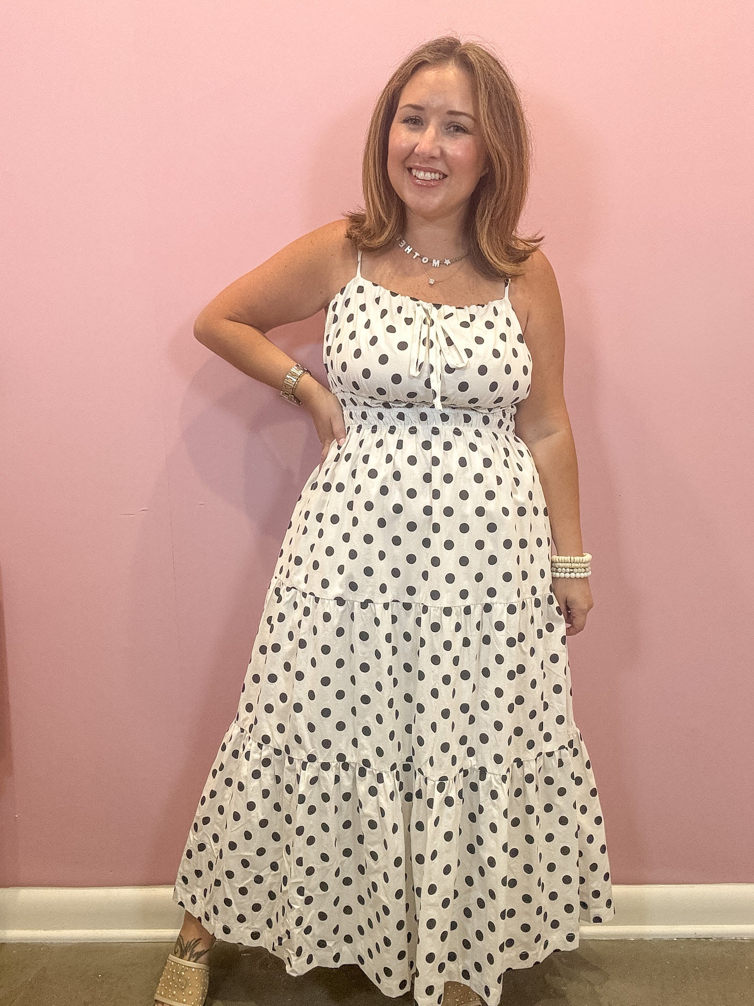 A woman modeling a white spaghetti-strap maxi dress with black polka dots, standing against a solid pink wall.