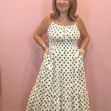 A woman modeling a white sleeveless maxi dress with black polka dots, standing against a solid pink wall.