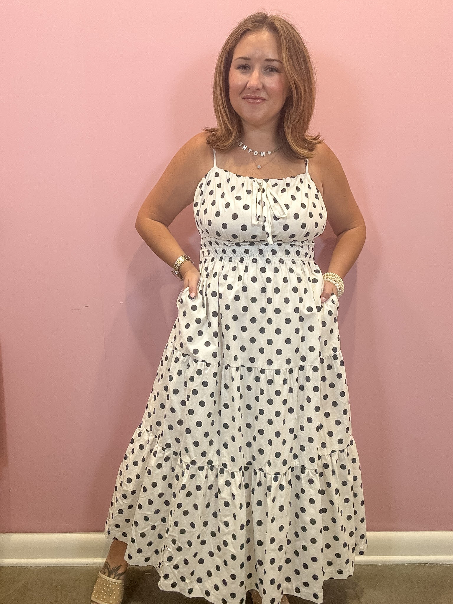 A woman modeling a white sleeveless maxi dress with black polka dots, standing against a solid pink wall.