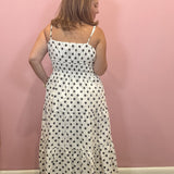 A woman wearing a white spaghetti-strap maxi dress with black polka dots, shown from the back against a solid pink wall.