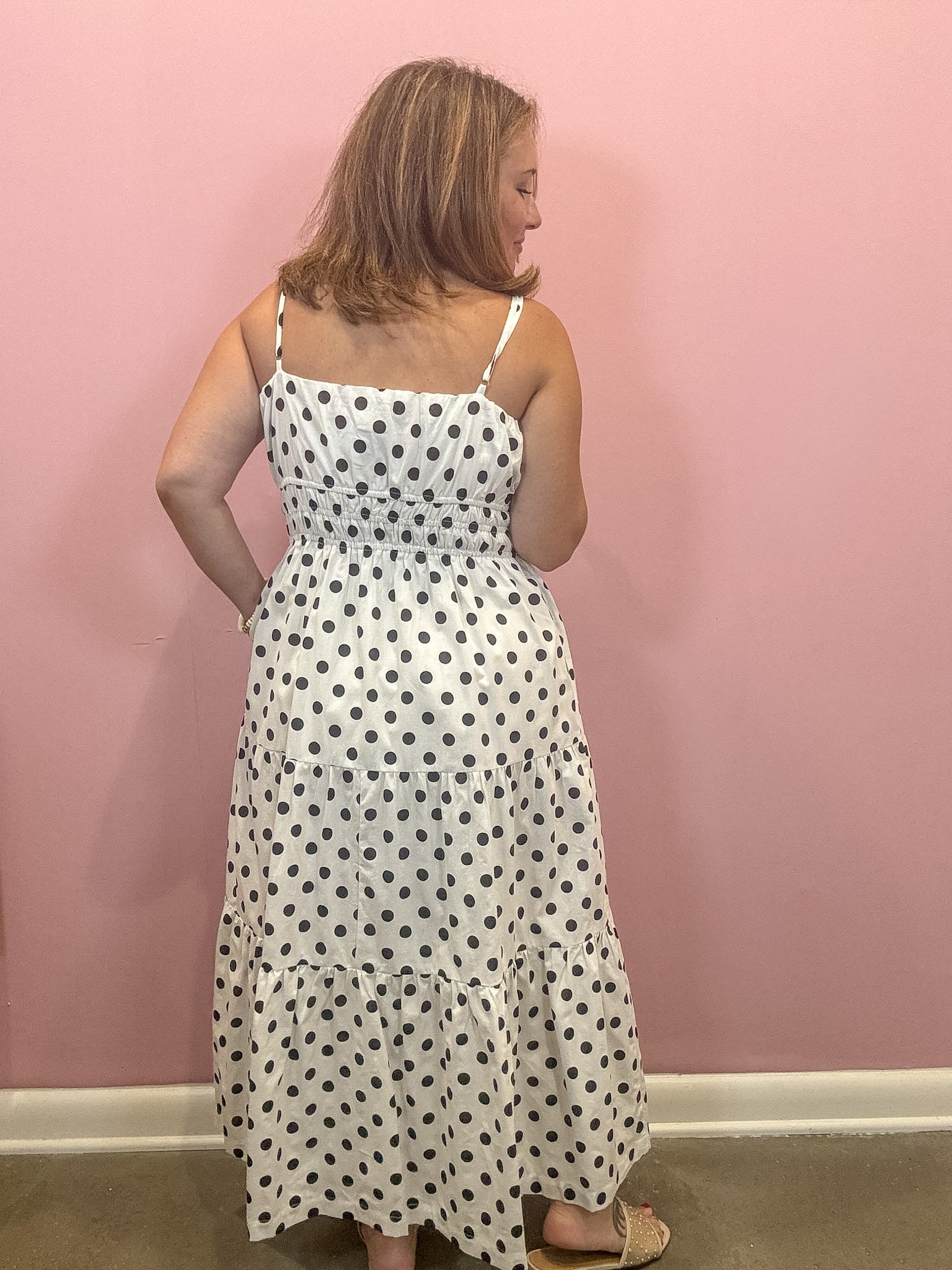A woman wearing a white spaghetti-strap maxi dress with black polka dots, shown from the back against a solid pink wall.