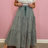 A person wearing a white short-sleeve button-front top and a long green-and-white gingham tiered skirt with a dark green drawstring, standing against a pink wall.