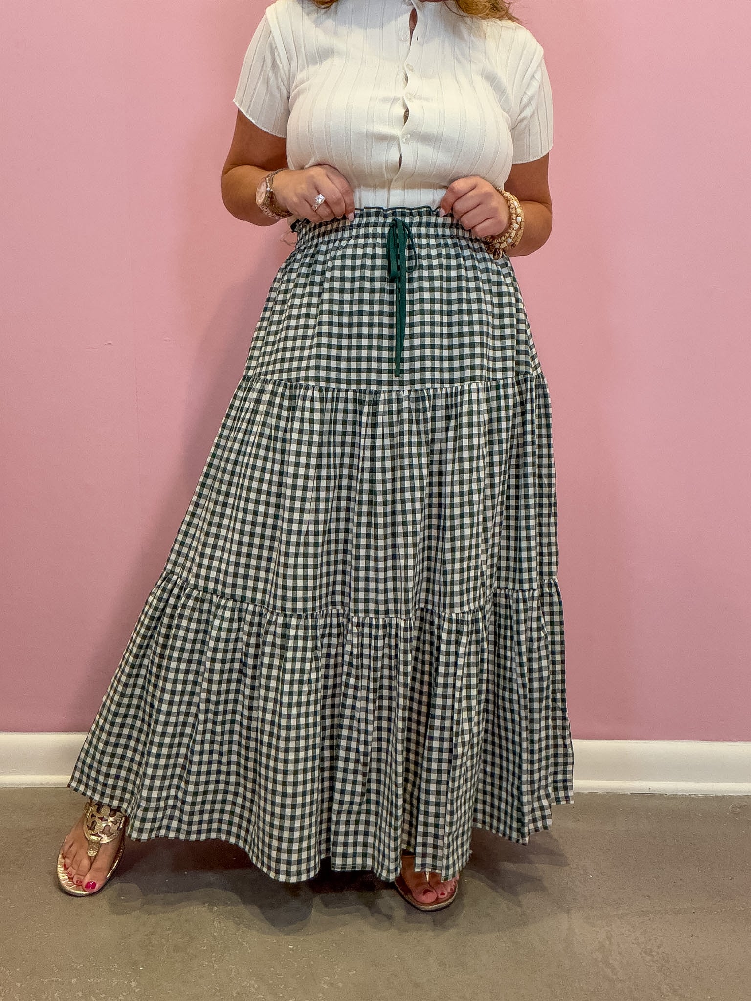 A person wearing a white short-sleeve button-front top and a long green-and-white gingham tiered skirt with a dark green drawstring, standing against a pink wall.