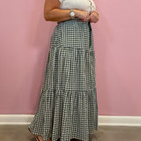 A woman wearing a white short-sleeve top and a black and white gingham tiered maxi skirt, standing against a solid pink wall.