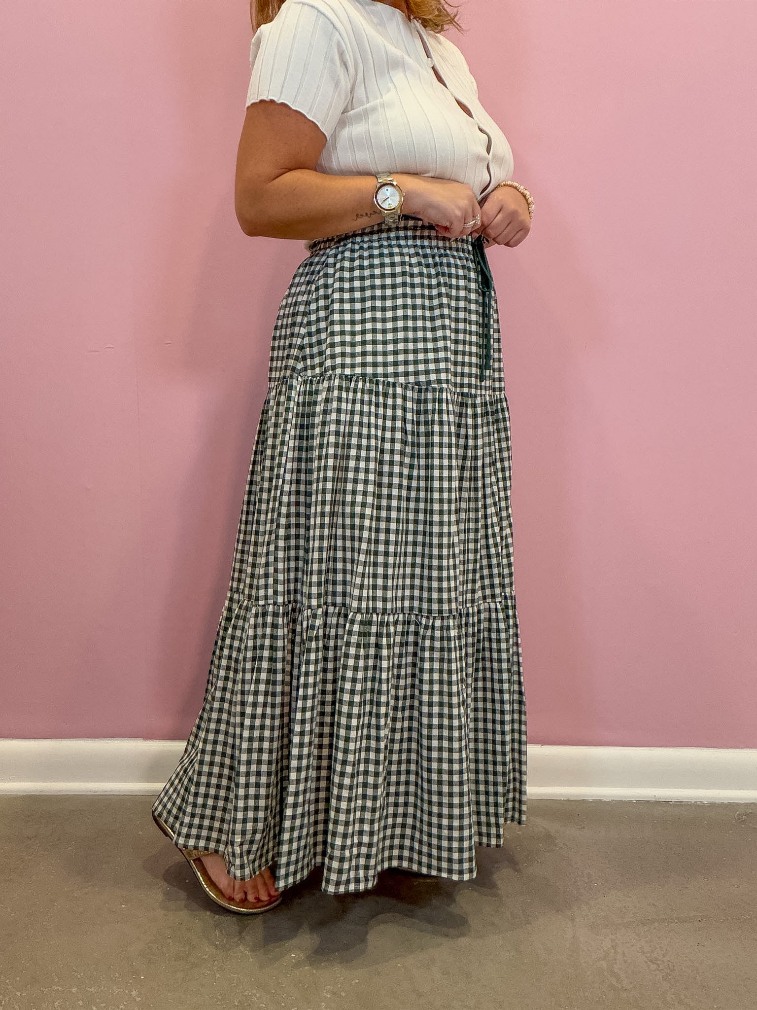 A woman wearing a white short-sleeve top and a black and white gingham tiered maxi skirt, standing against a solid pink wall.