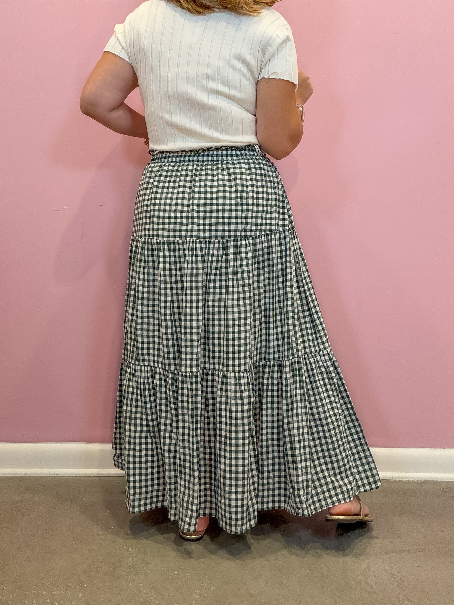 A woman shown from the back wearing a long tiered gingham maxi skirt in green and white, paired with a white ribbed short-sleeve top, standing against a pink wall.
