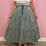 A person wearing a white short-sleeve button-front top and a long green-and-white gingham tiered skirt with a dark green drawstring, standing against a pink wall.