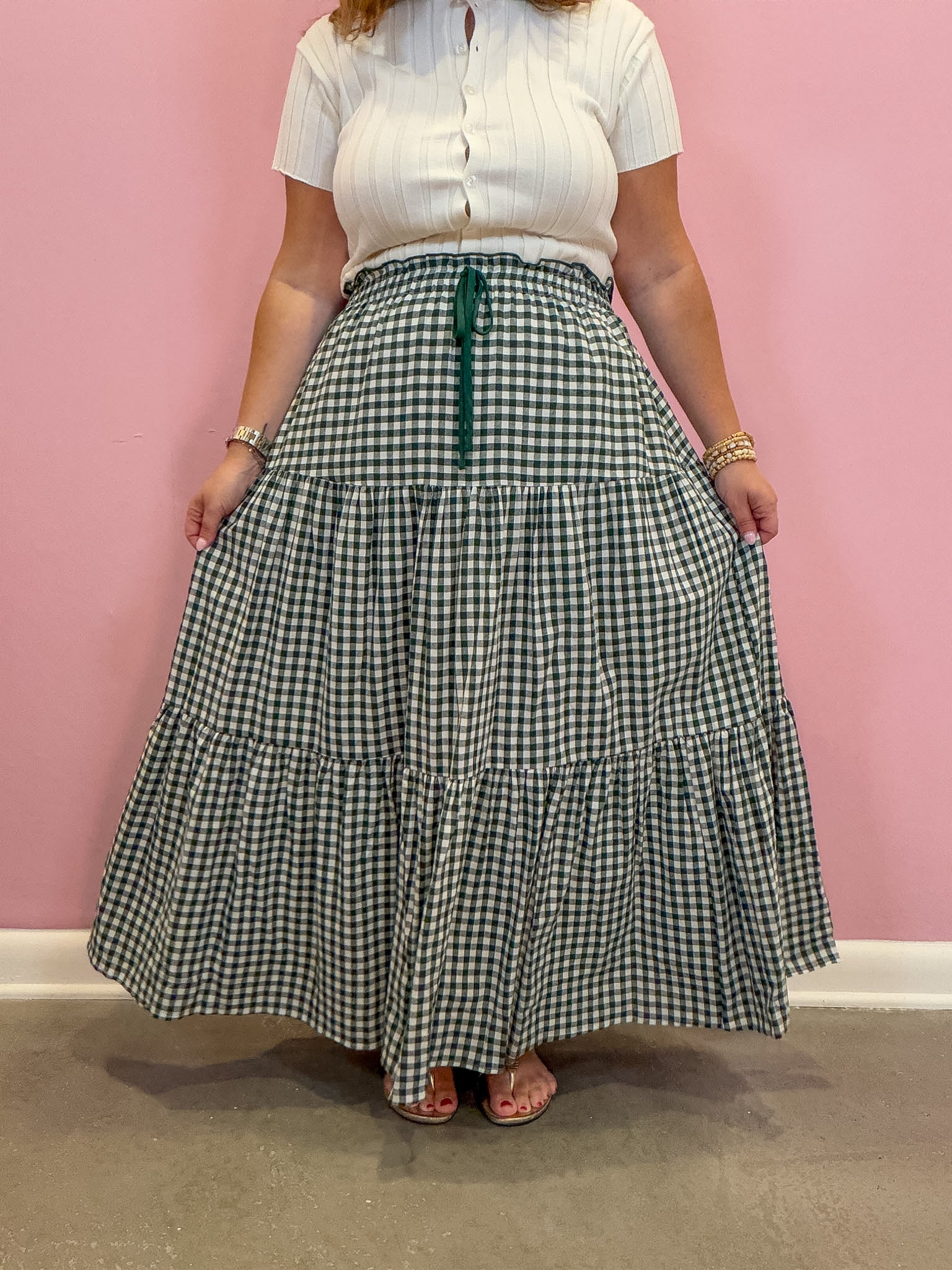 A person wearing a white short-sleeve button-front top and a long green-and-white gingham tiered skirt with a dark green drawstring, standing against a pink wall.