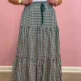 A person wearing a long tiered gingham maxi skirt in green and white with a dark green drawstring waist, paired with a white ribbed top, standing against a pink wall.