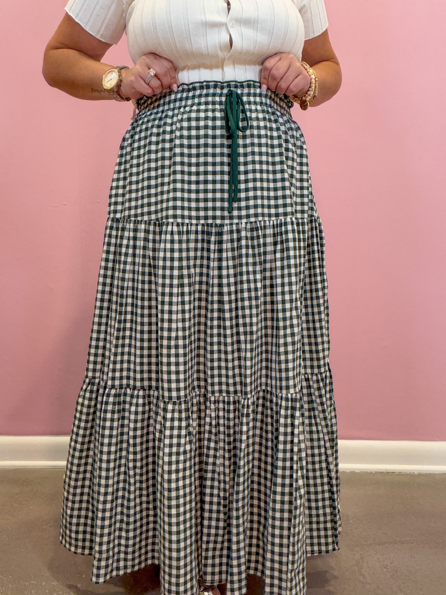 A person wearing a long tiered gingham maxi skirt in green and white with a dark green drawstring waist, paired with a white ribbed top, standing against a pink wall.