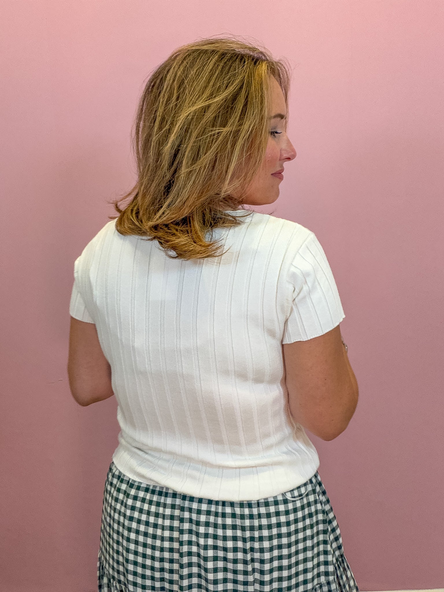 A woman with blonde hair is shown from the back wearing a white short-sleeve ribbed mock neck top and a green and white gingham skirt, standing against a solid pink wall.