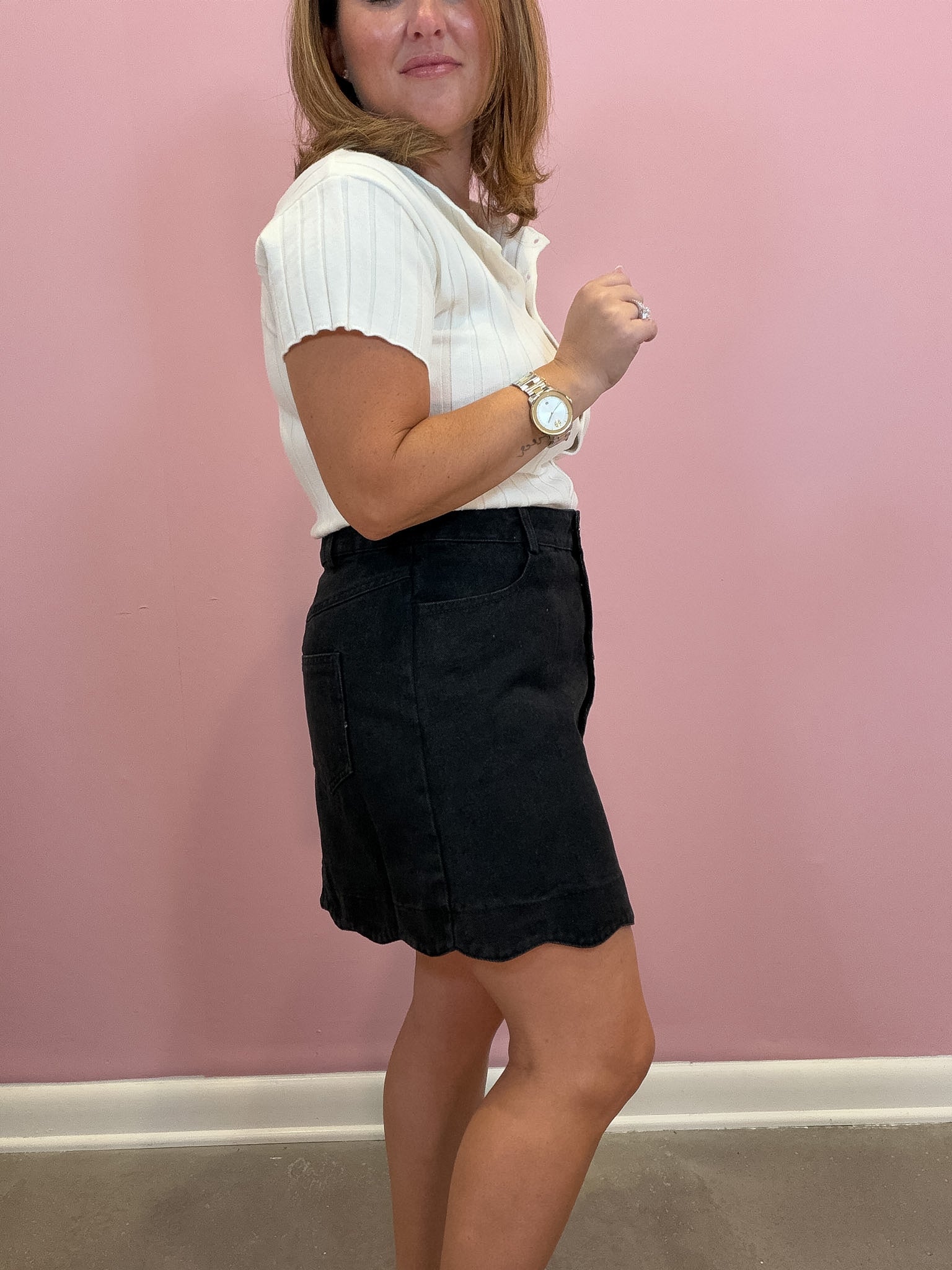 A woman modeling a white short-sleeve ribbed knit top and a black denim mini skirt against a pink wall.