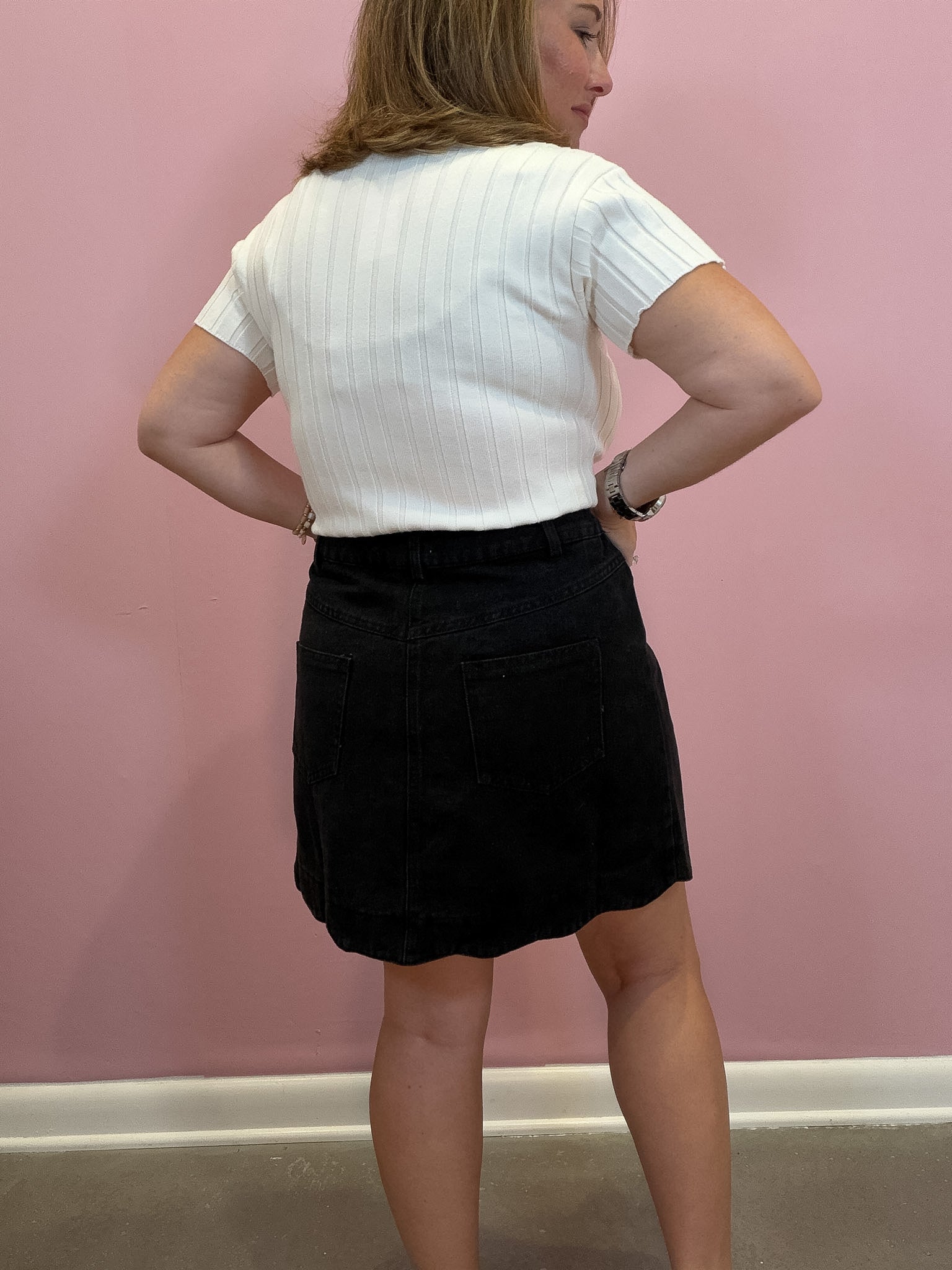 A woman is shown from the back wearing a white short-sleeve ribbed knit top and a black corduroy mini skirt, standing against a solid pink wall.
