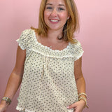 A smiling woman wearing a cream-colored two-piece pajama set with a small gray floral print, featuring a ruffled neckline and ruffled hem, posed against a solid pink wall.