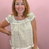 A smiling woman with light brown hair stands against a solid pink wall wearing a matching cream-colored two-piece pajama set with a small gray floral print and white ruffle trim.