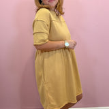 A woman modeling a mustard yellow short-sleeve dress against a pink wall.