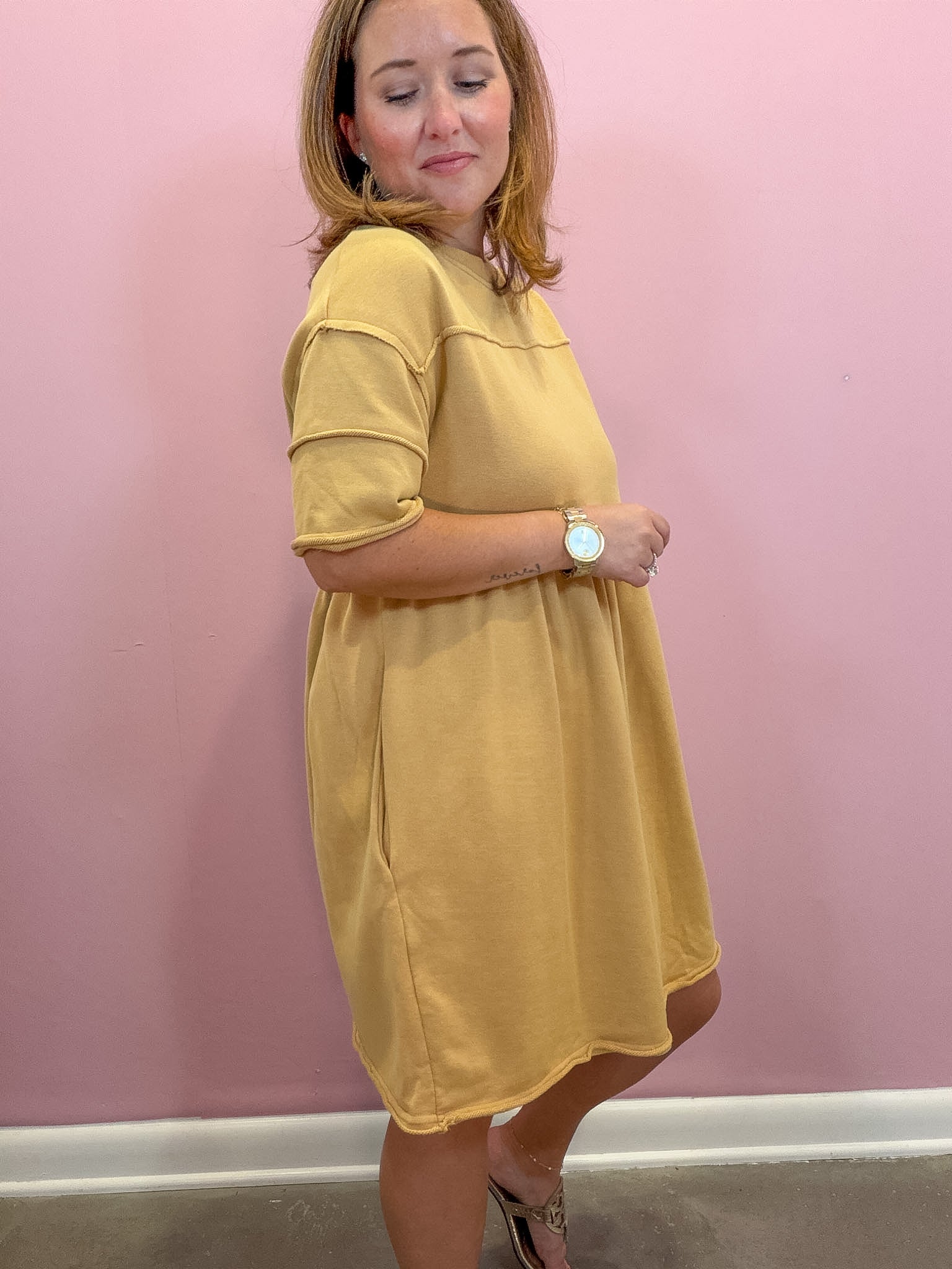 A woman modeling a mustard yellow short-sleeve dress against a pink wall.