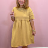 A woman modeling a mustard yellow short-sleeve dress with a gathered waist, standing against a solid pink wall.