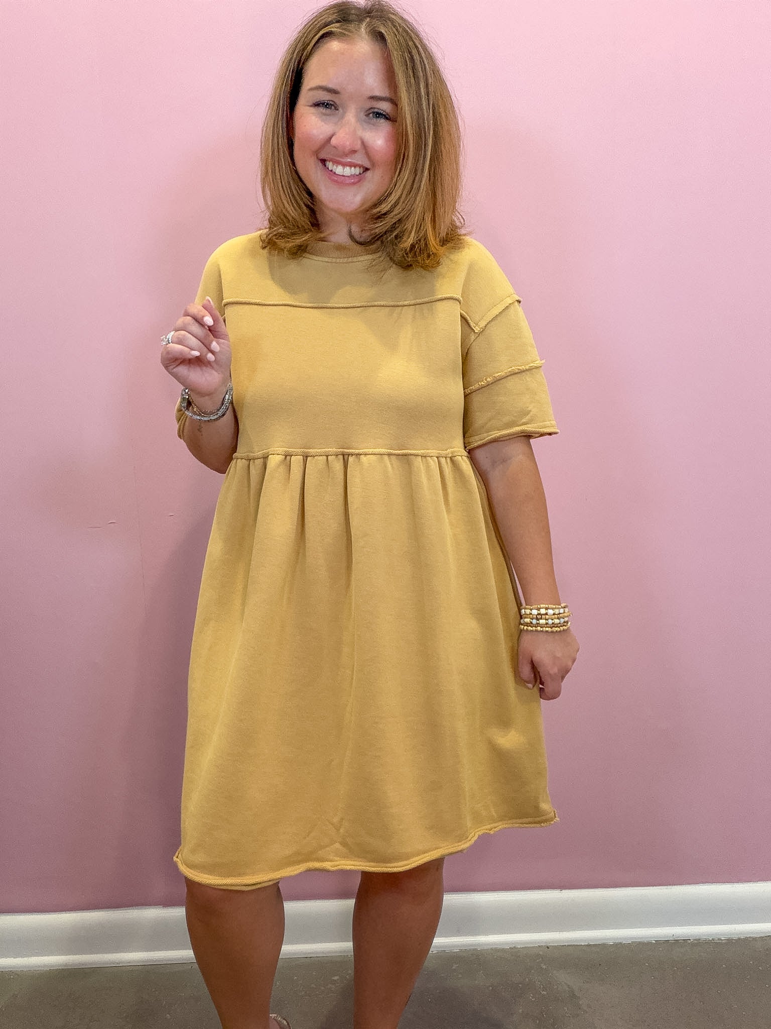 A woman modeling a mustard yellow short-sleeve dress with a gathered waist, standing against a solid pink wall.