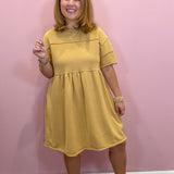 A woman modeling a mustard yellow short-sleeve dress with a gathered waist, standing against a solid pink wall.