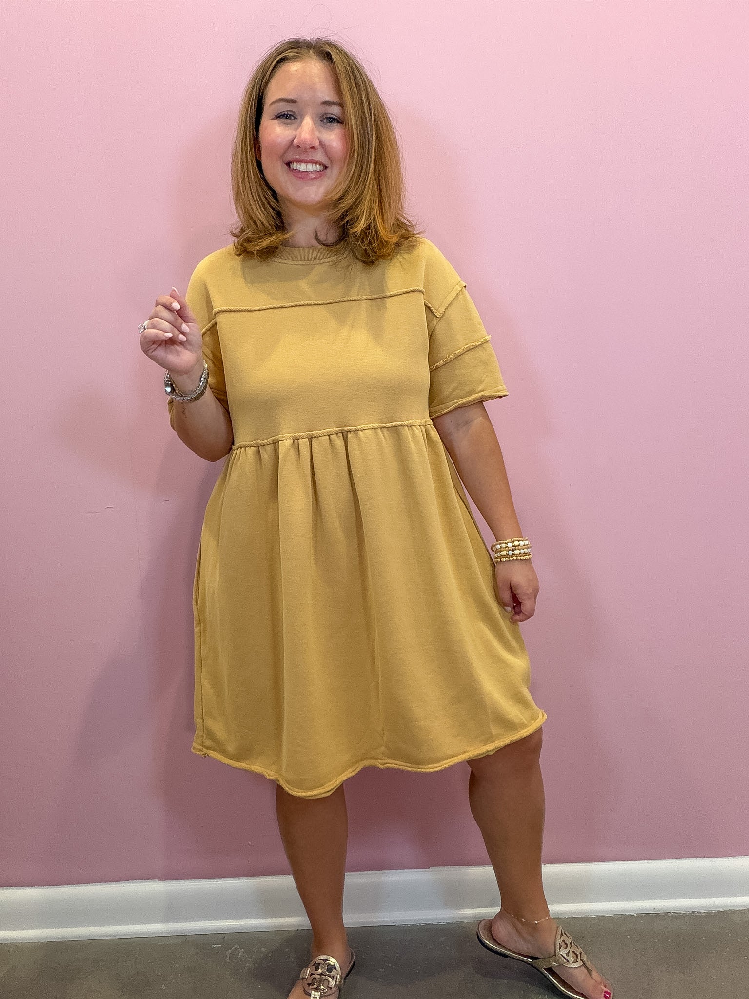 A woman modeling a mustard yellow short-sleeve dress with a gathered waist, standing against a solid pink wall.