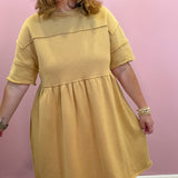 A woman wearing a mustard yellow short-sleeve dress with a gathered waist, standing against a solid pink wall.