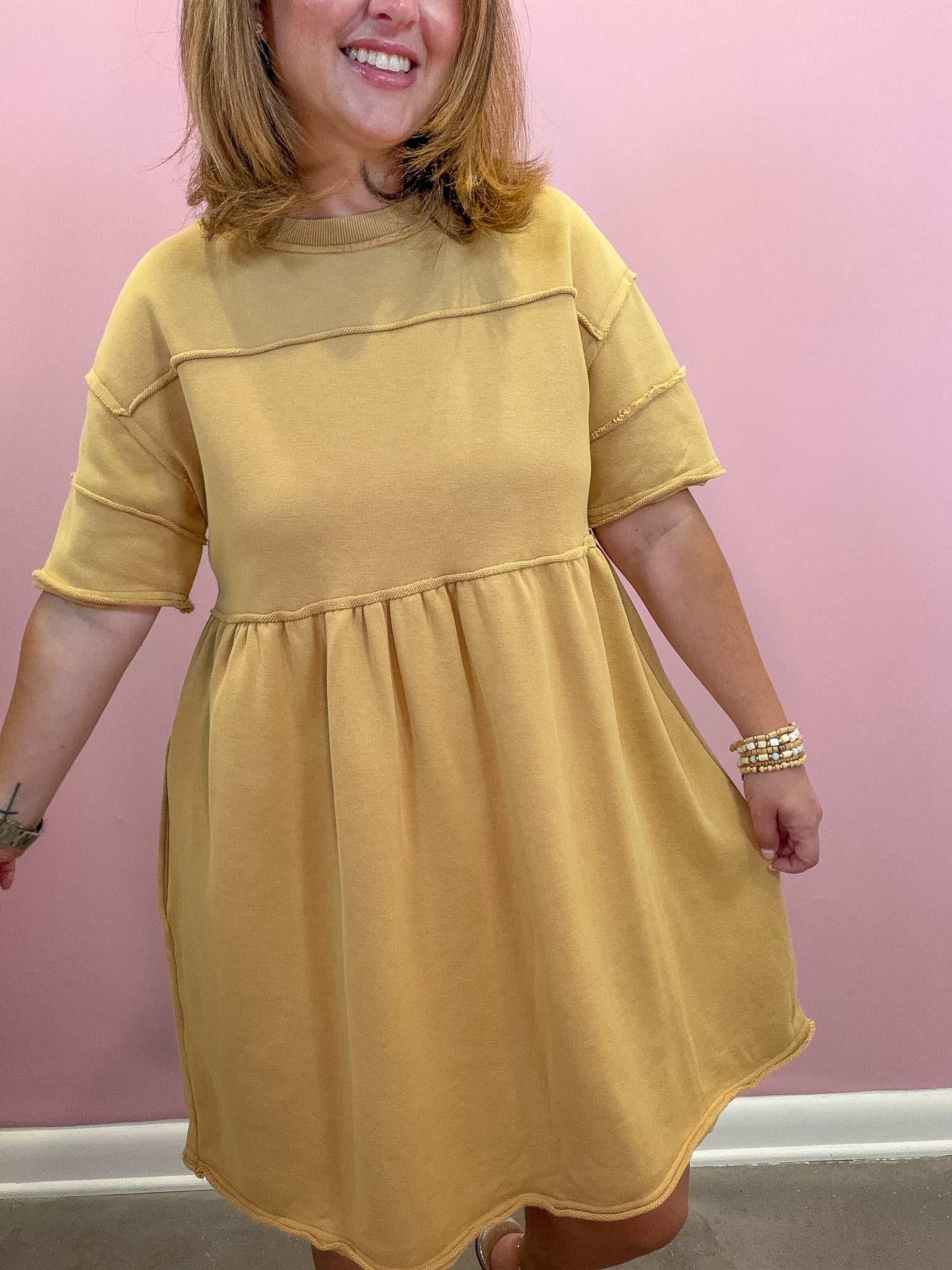 A woman wearing a mustard yellow short-sleeve dress with a gathered waist, standing against a solid pink wall.