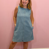 A woman wearing a sleeveless light blue corduroy dress with side pockets, standing against a solid pink wall.
