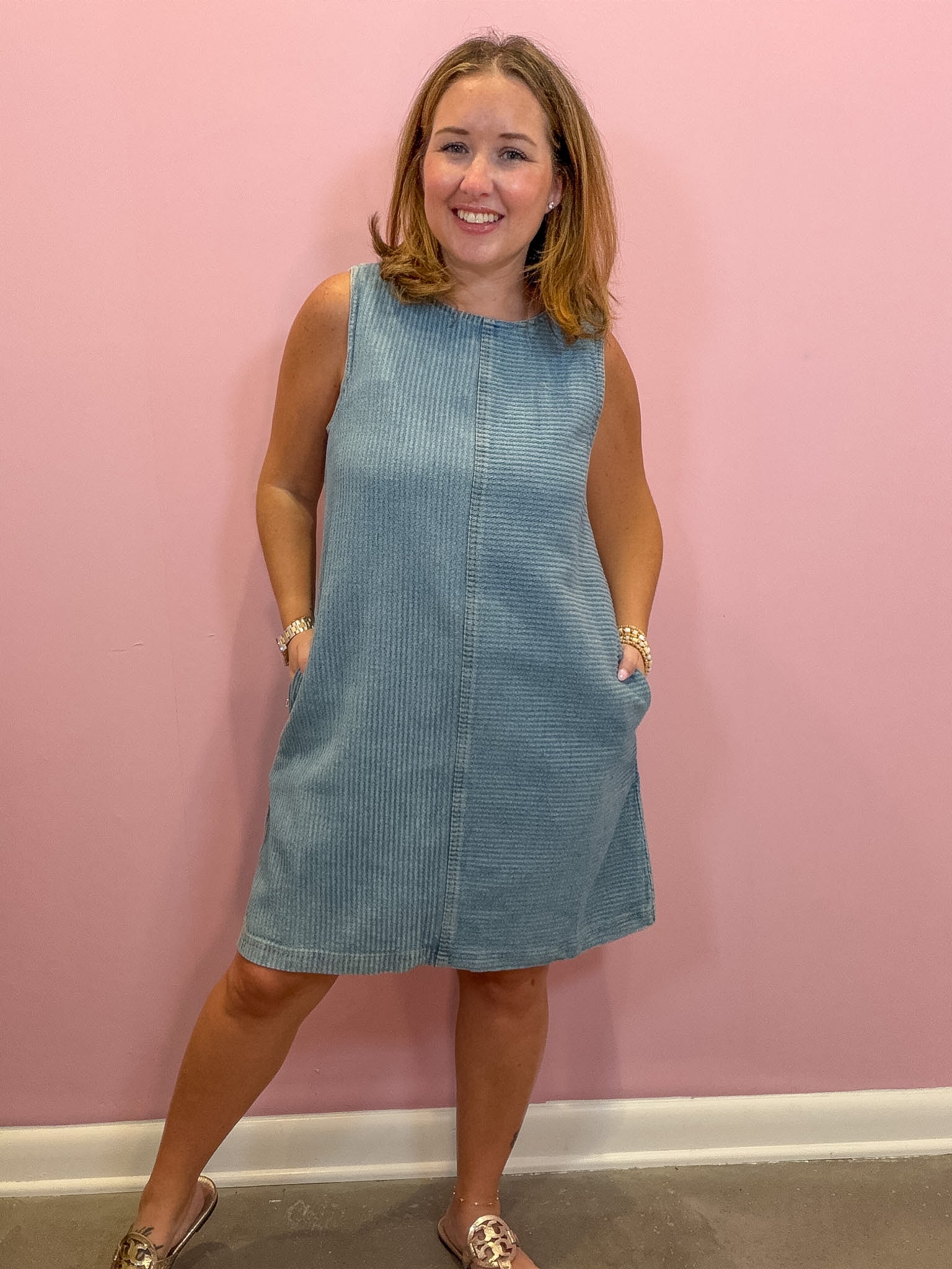 A woman wearing a sleeveless light blue corduroy dress with side pockets, standing against a solid pink wall.