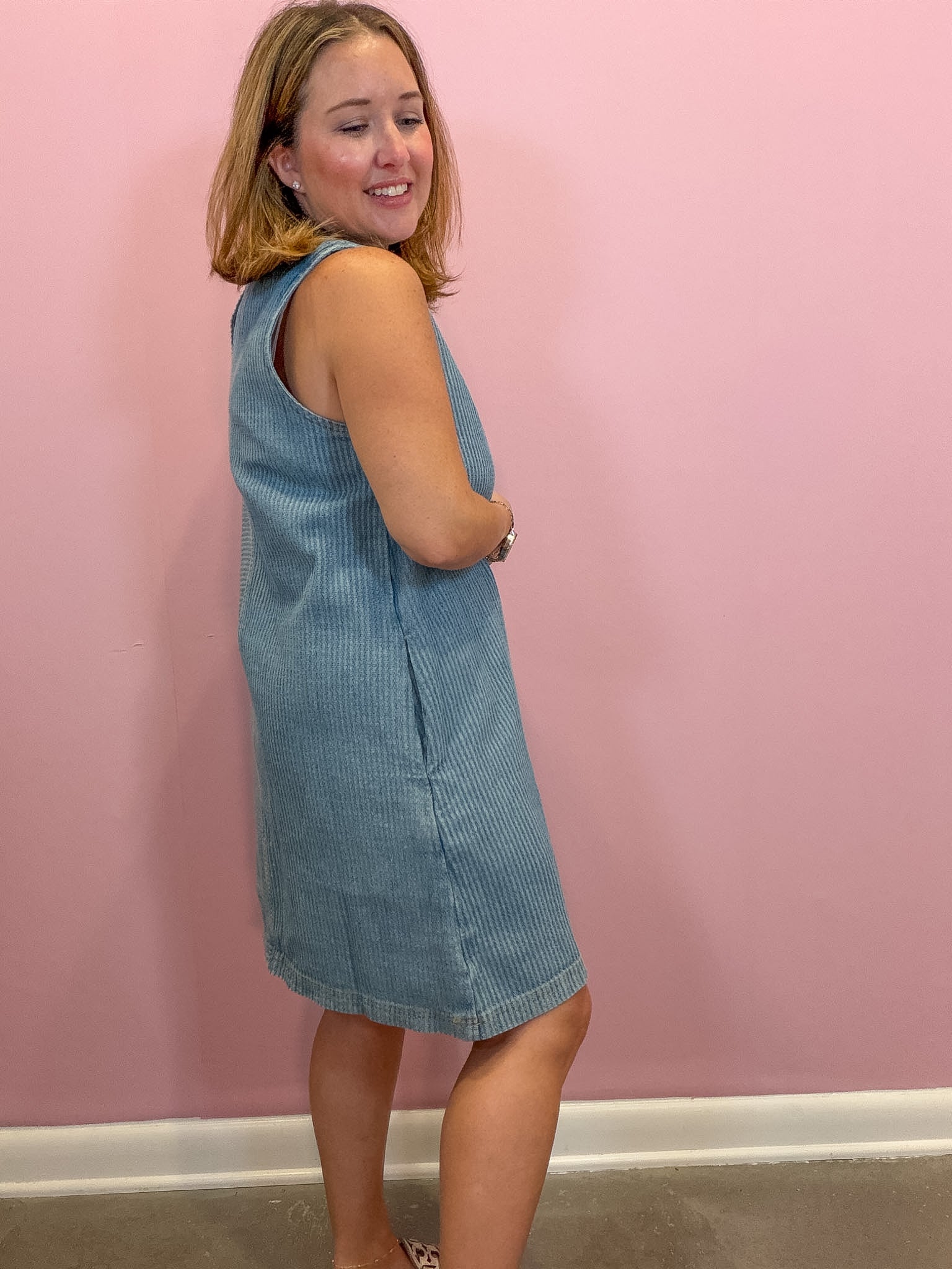 A woman modeling a light blue corduroy sleeveless dress against a solid pink wall.